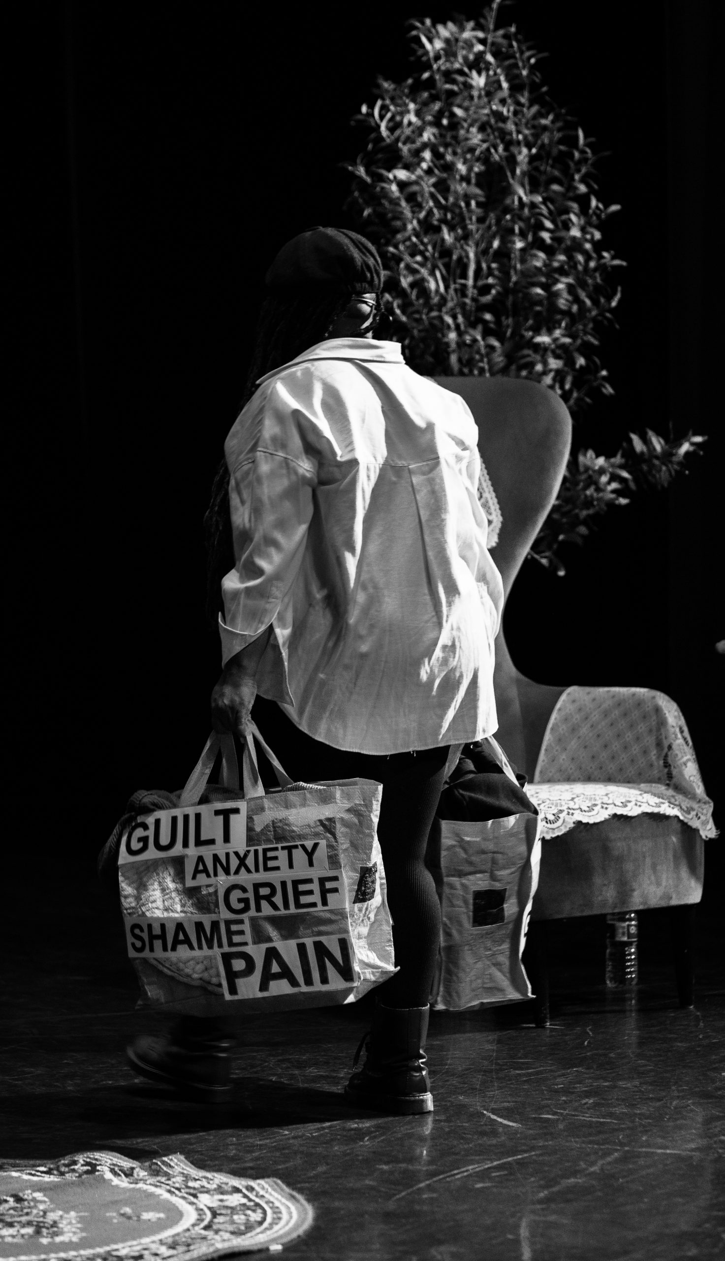 a B&W image of Yvonne taken at the shaw theatre in may 2026, Caught You in a Lie, in the image Yvonne is carrying a bag that externally shows all the negative thoughts we have about ourselves